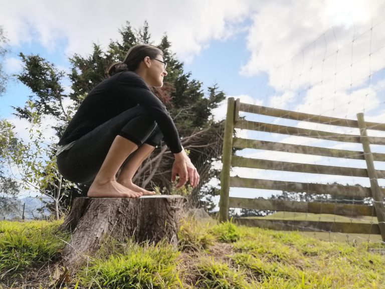 emma dressed in black squatting on a tree log in a field against a blue sky with a wooden fence in the background.