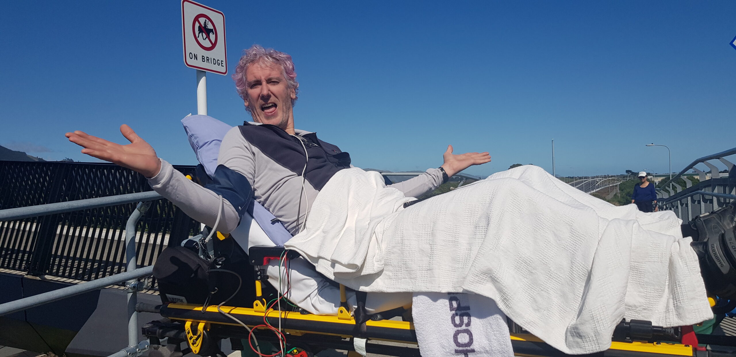 man on a gurney type stretcher with arms outstretched laughing against a blue sky.